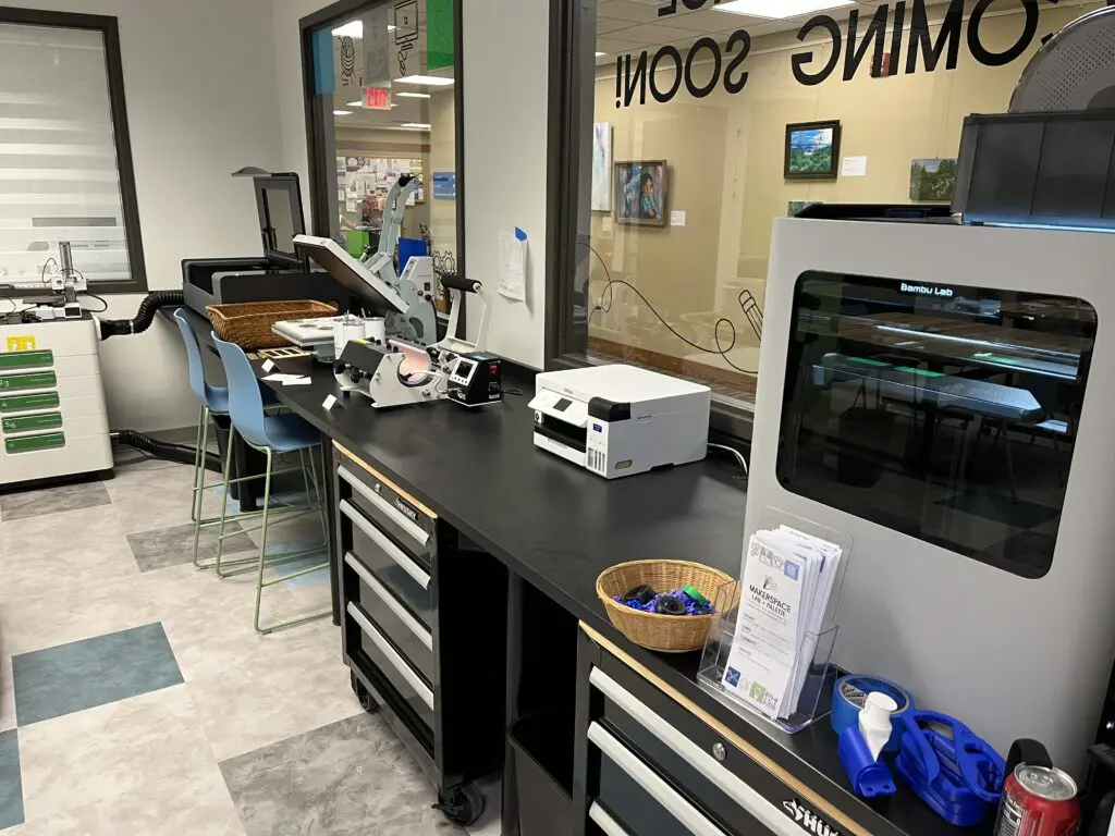 MakerspaceLabMachines A counter spanning the length of a wall. On top of the counter sits a laser engraver, clamshell and mug heat presses, a sublimation printer, and a 3D printer.