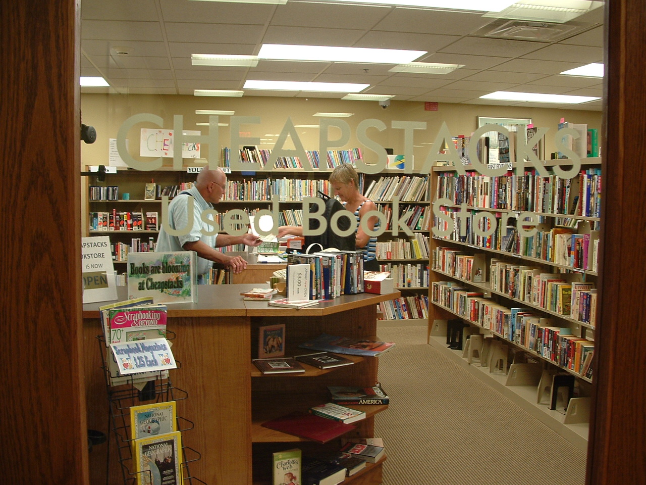 Image of Cheapstacks Used Bookstore front door. Bookshelves, a patron checking out a book, and a volunteer assisting the patron can be seen through the door's glass.