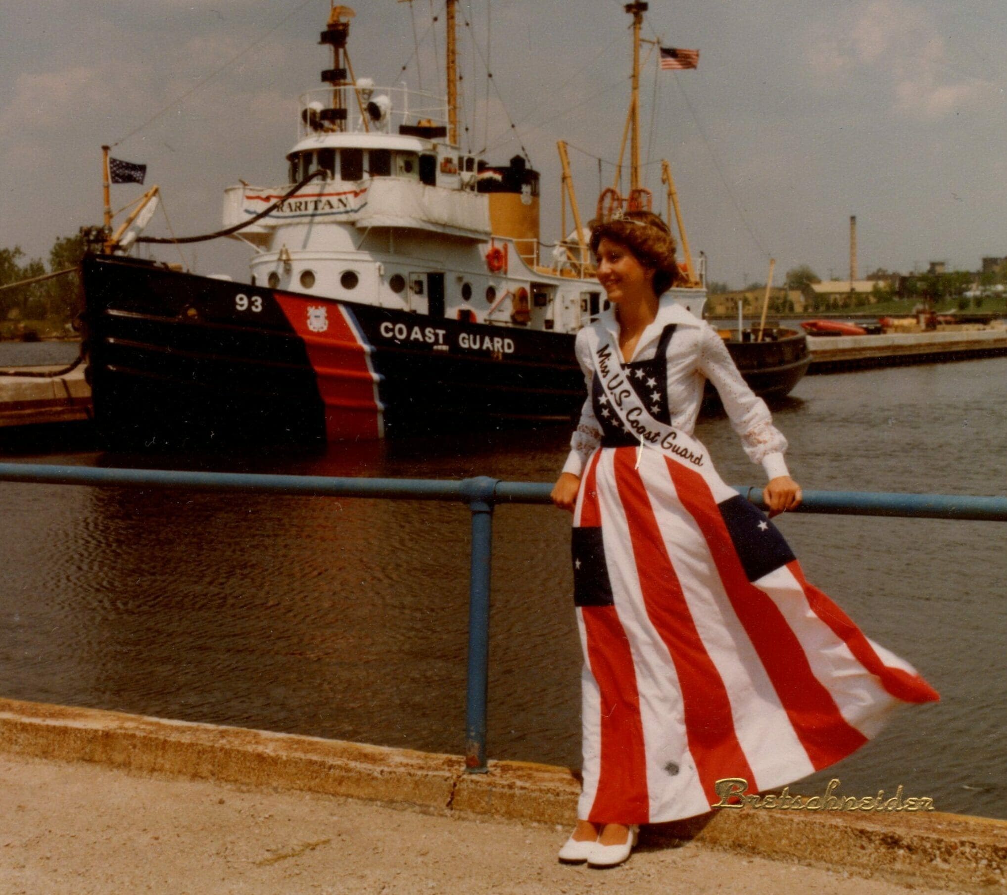 dated photograph of Miss U.S. Coast Guard standing at the edge of the waterfront in front of a Coast Guard vessel and smiling