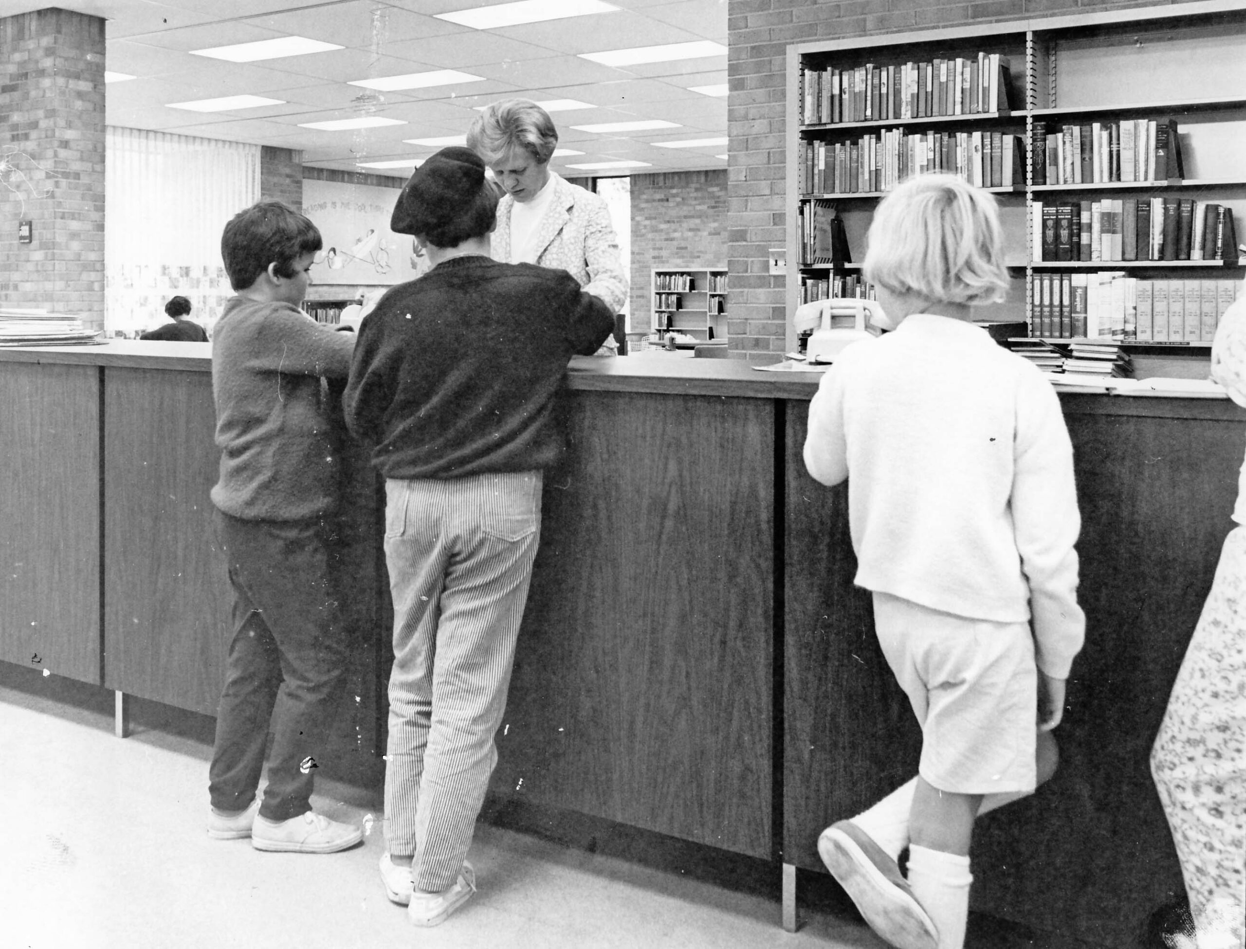Black and White photograph of well-dressed children at a counter talking with a librarian at an old library